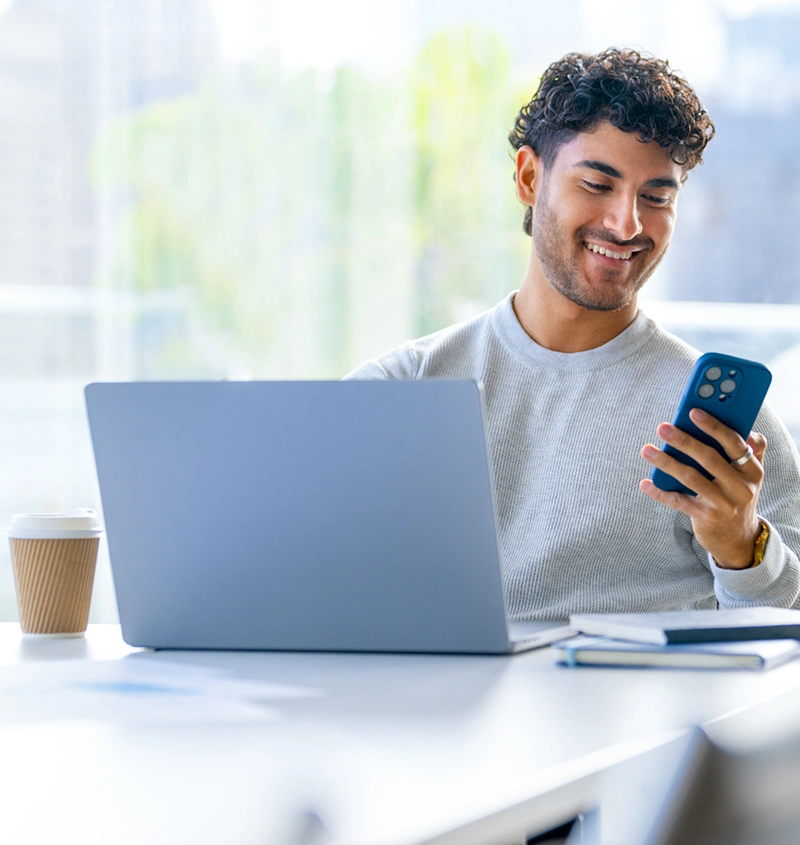 A young man with curly hair smiles while looking at his blue phone, sitting by a laptop.