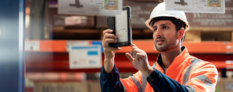 Warehouse worker wearing a hard hat and high-visibility jacket using a tablet in a distribution center