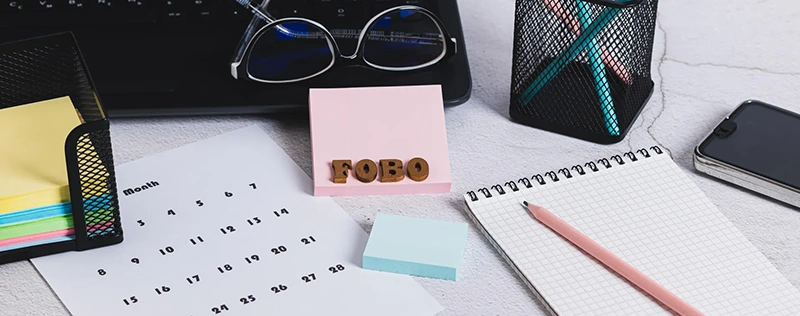 Office desk with calendar, notebook, sticky notes, and a note spelling “FOBO”